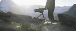 Portrait d’un photographe en montagne, observant au loin avec des jumelles, sur fond de sommets enneigés.